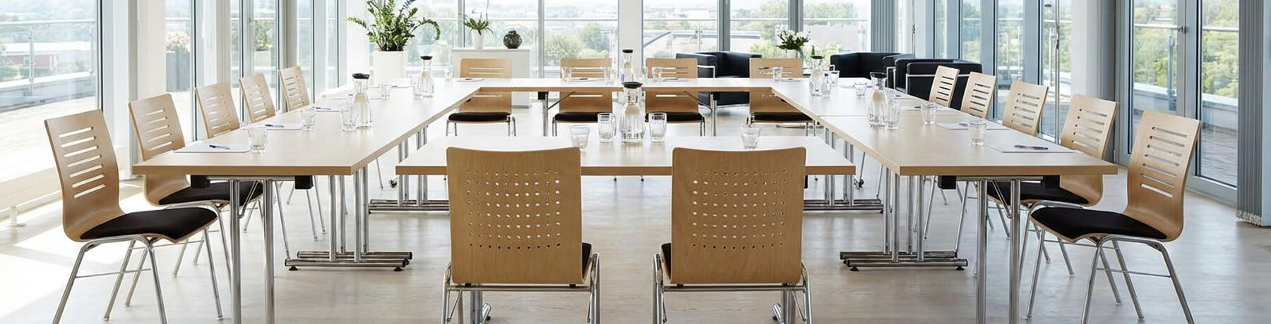 Header_teaser_konferenzmoebel Heller, moderner Konferenzraum mit U-förmig angeordneten Tischen und stapelbaren Holzstühlen – professionelle Konferenzmöbel für Meetings und Seminare.