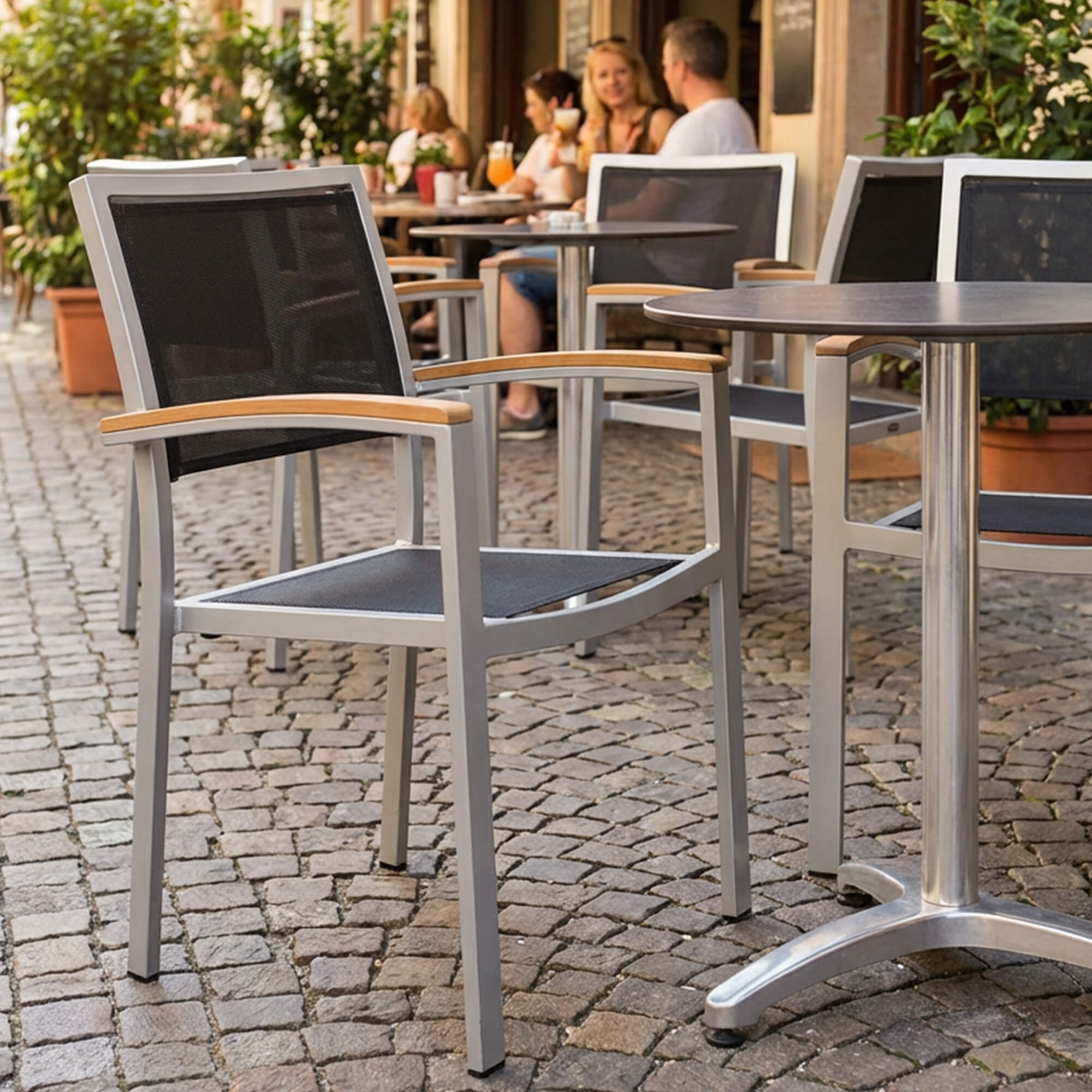 Outdoorstuhl Georgia – silber-schwarzer Outdoorstuhl mit Armlehnen vor Caféterrasse auf Kopfsteinpflaster