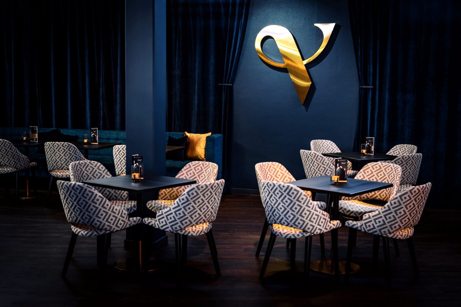 Lounge-Sitzgruppe mit Logo im Hintergrund Lounge-Sitzgruppe im Zaubertheater VERSCHMITTST mit gemusterten Stühlen, kleinen Tischen und Logo im Hintergrund
